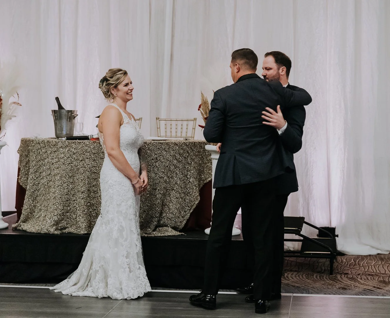 A bride and two grooms during a wedding ceremony, with one groom hugging the other as the bride watches and smiles.