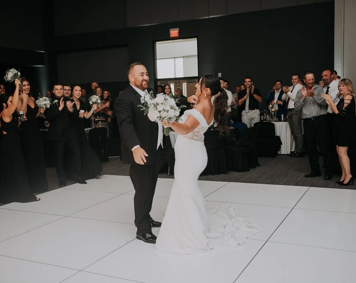 Bride and groom dancing during their wedding reception, surrounded by smiling guests clapping and cheering in a dimly lit indoor venue.