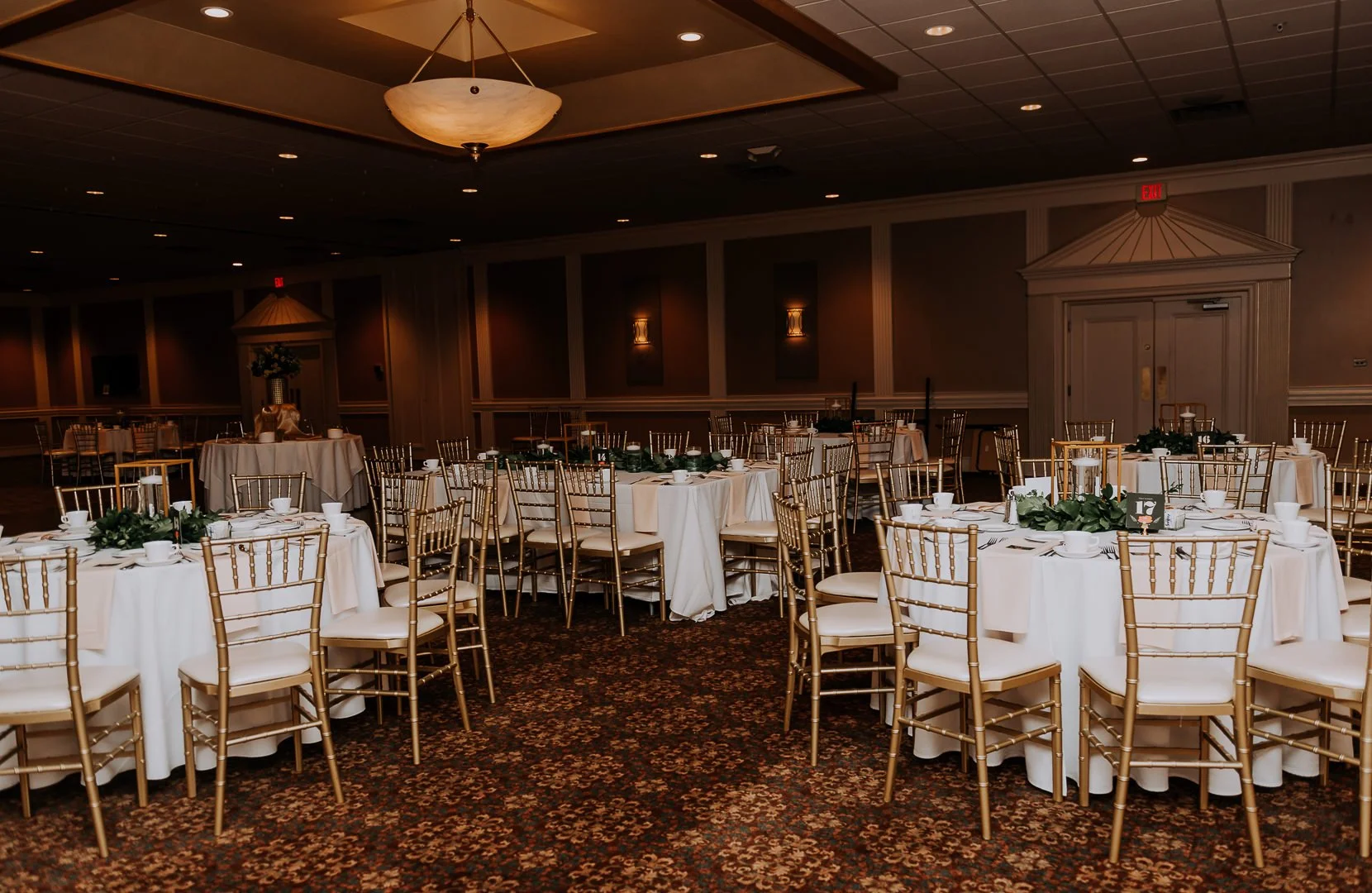 Elegant banquet hall with round tables covered in white tablecloths, gold chairs, and floral centerpieces, set for a formal event.