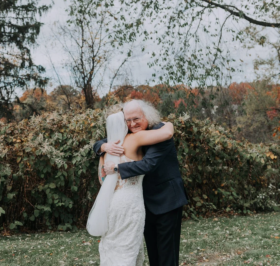 Older man and young woman hugging outdoors, surrounded by fall foliage.