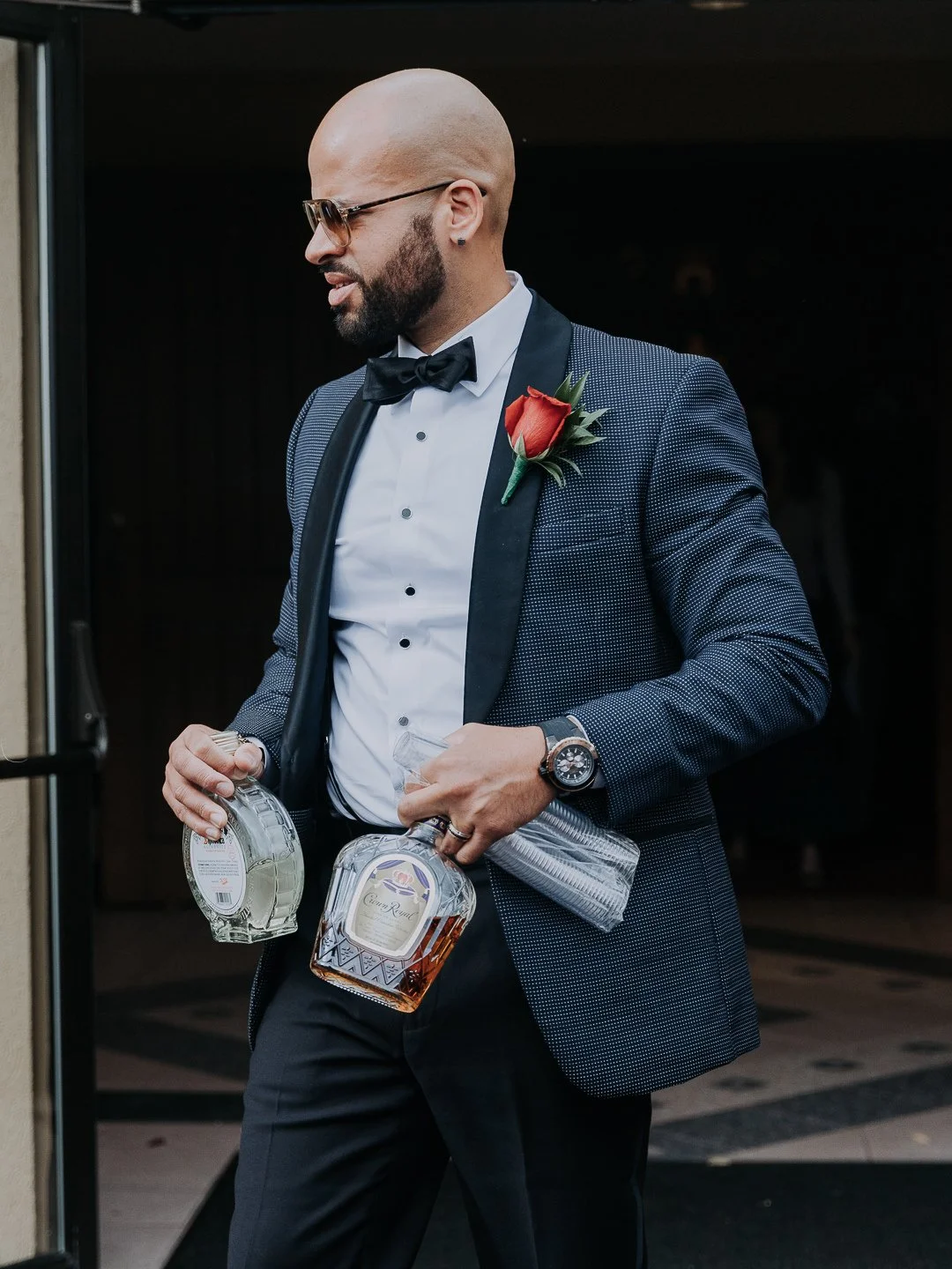 A man wearing a tuxedo with a red boutonniere, glasses, and a wristwatch, holding two bottles of liquor, standing outside a building.