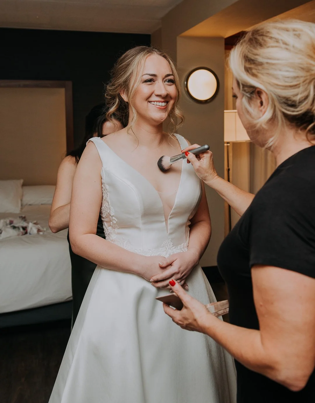 A bride is smiling while getting her makeup done by a makeup artist in a hotel room.