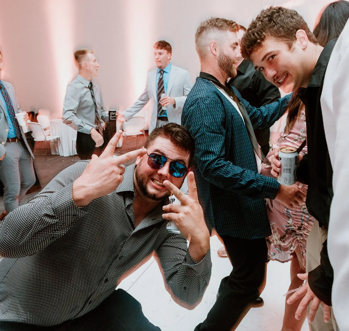 Group of young adults at a party, socializing and having fun. Some are holding drinks, smiling, and making hand gestures.