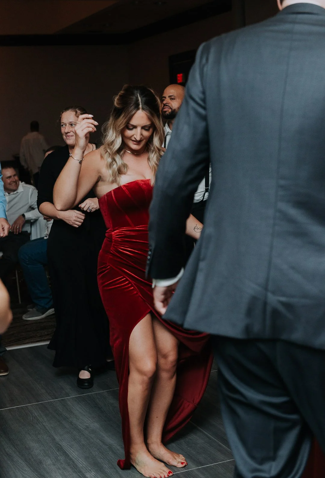 A woman in a red strapless evening gown with a thigh-high slit dancing barefoot at an indoor event, with other guests seated and standing around her, some smiling and watching.