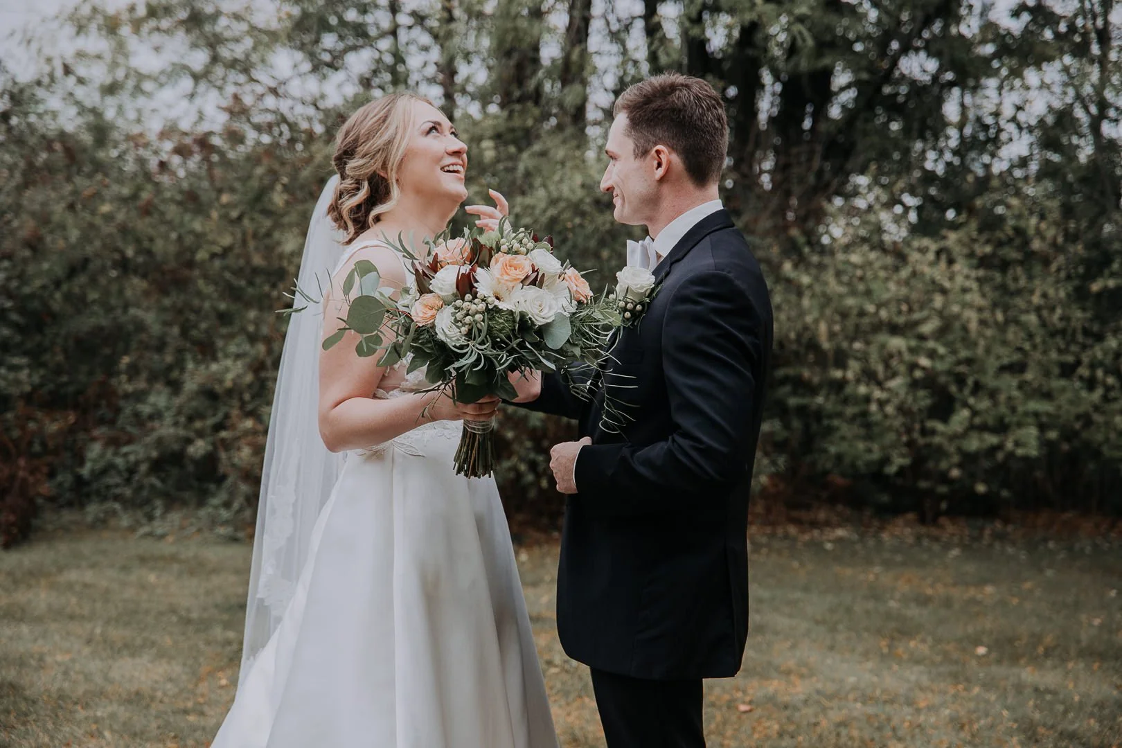 A bride and groom sharing a joyful moment with the bride holding a bouquet of flowers outdoors, surrounded by trees.