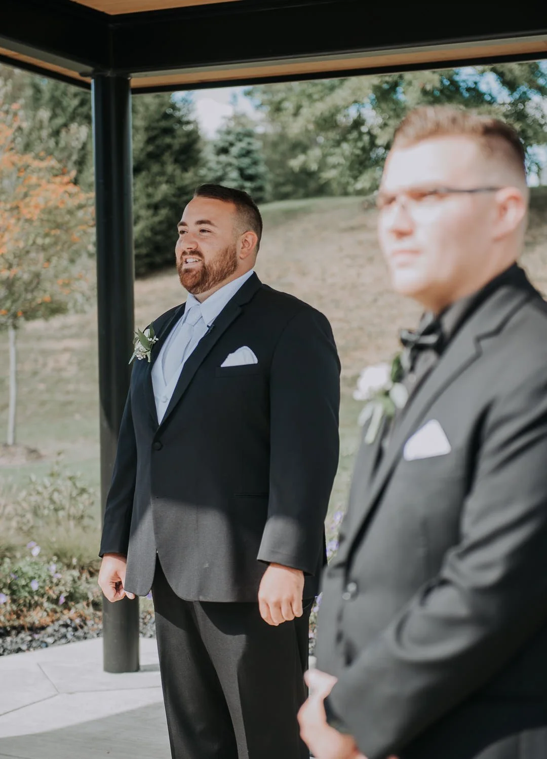 Two men dressed in tuxedos at an outdoor wedding ceremony, standing in front of a lush green landscape with trees.