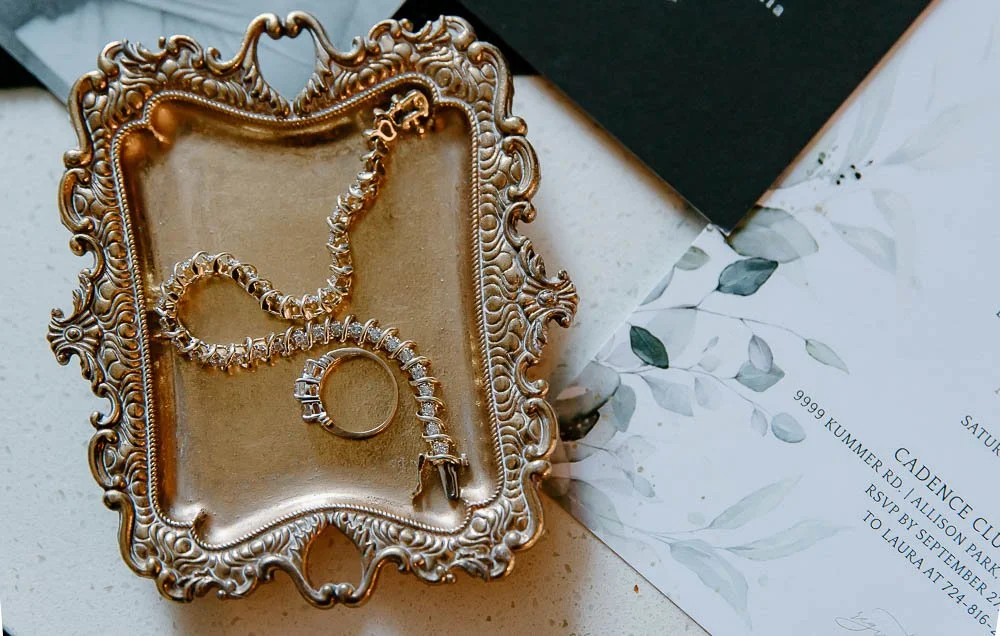 Silver jewelry including a necklace, bracelet, and ring on an ornate gold tray, with a floral-themed invitation or card next to it.