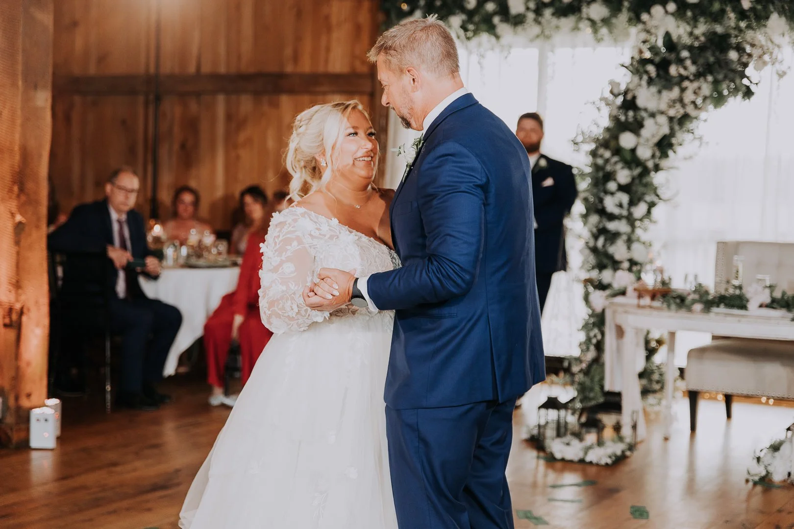 A bride and groom share a dance during their wedding reception. The bride is wearing a white lace wedding gown, and the groom is dressed in a navy blue suit. They are smiling at each other while holding hands.