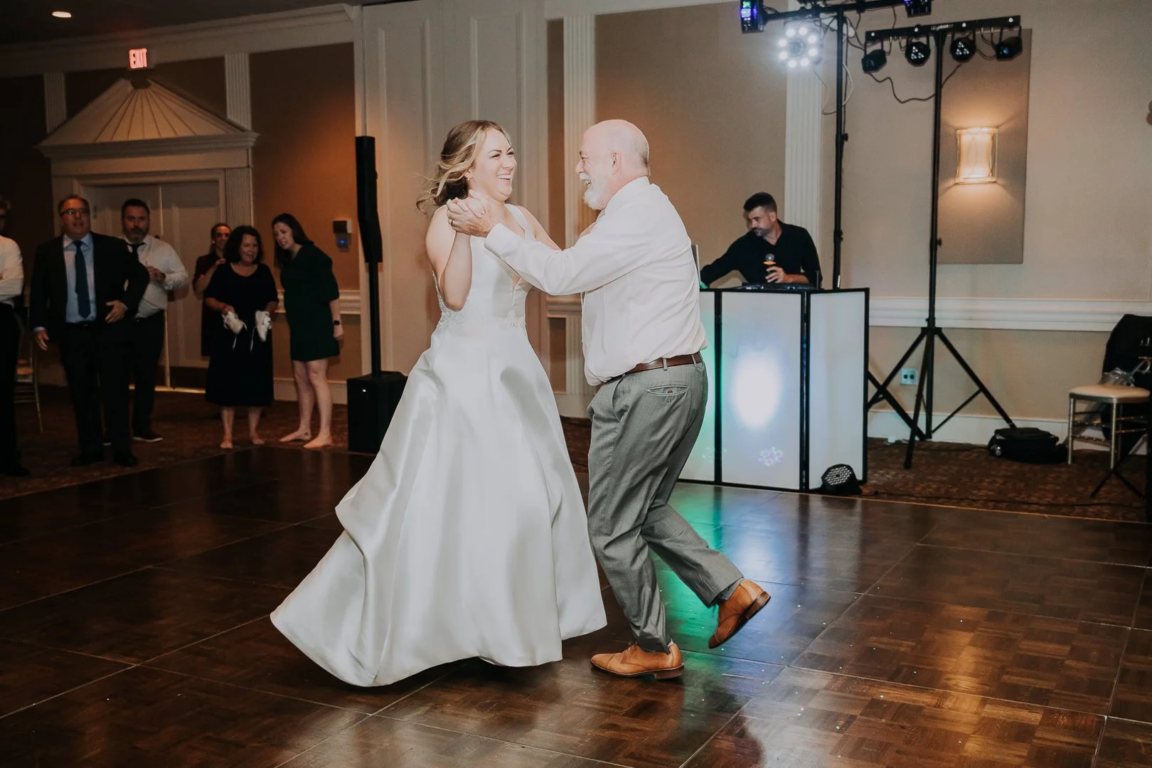 A bride and an older man dancing together at a wedding reception, surrounded by guests and a DJ setup in the background.
