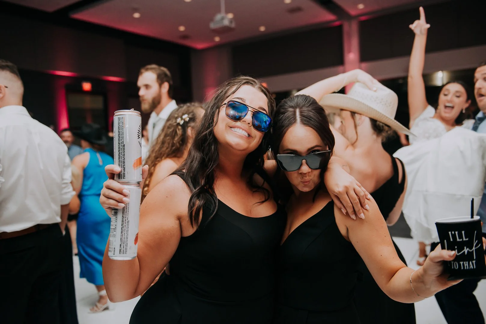 Two women in black dresses wearing sunglasses, smiling and dancing at a lively party or celebration.