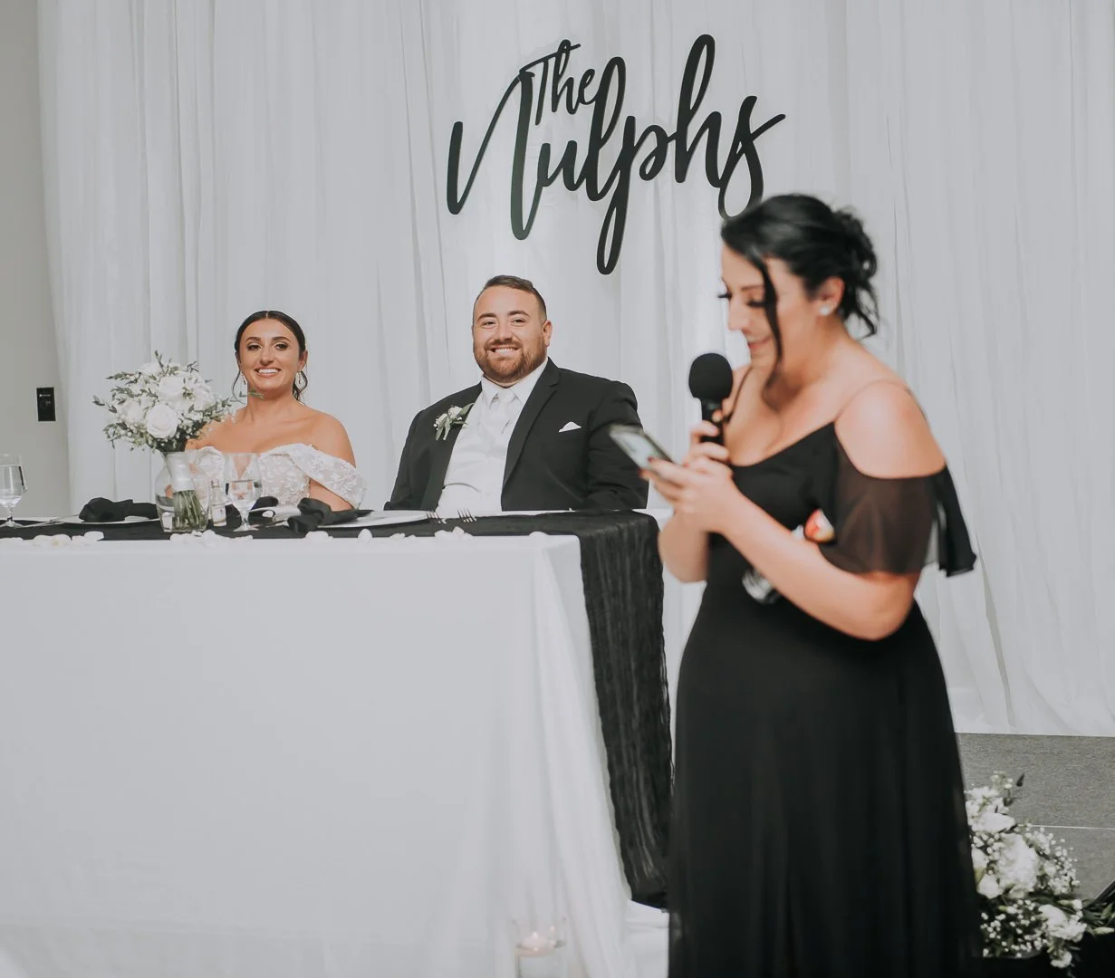 A wedding reception scene with a bride and groom sitting at a table and a woman giving a speech with a microphone. The backdrop has the word 'The Nulphs' in cursive.