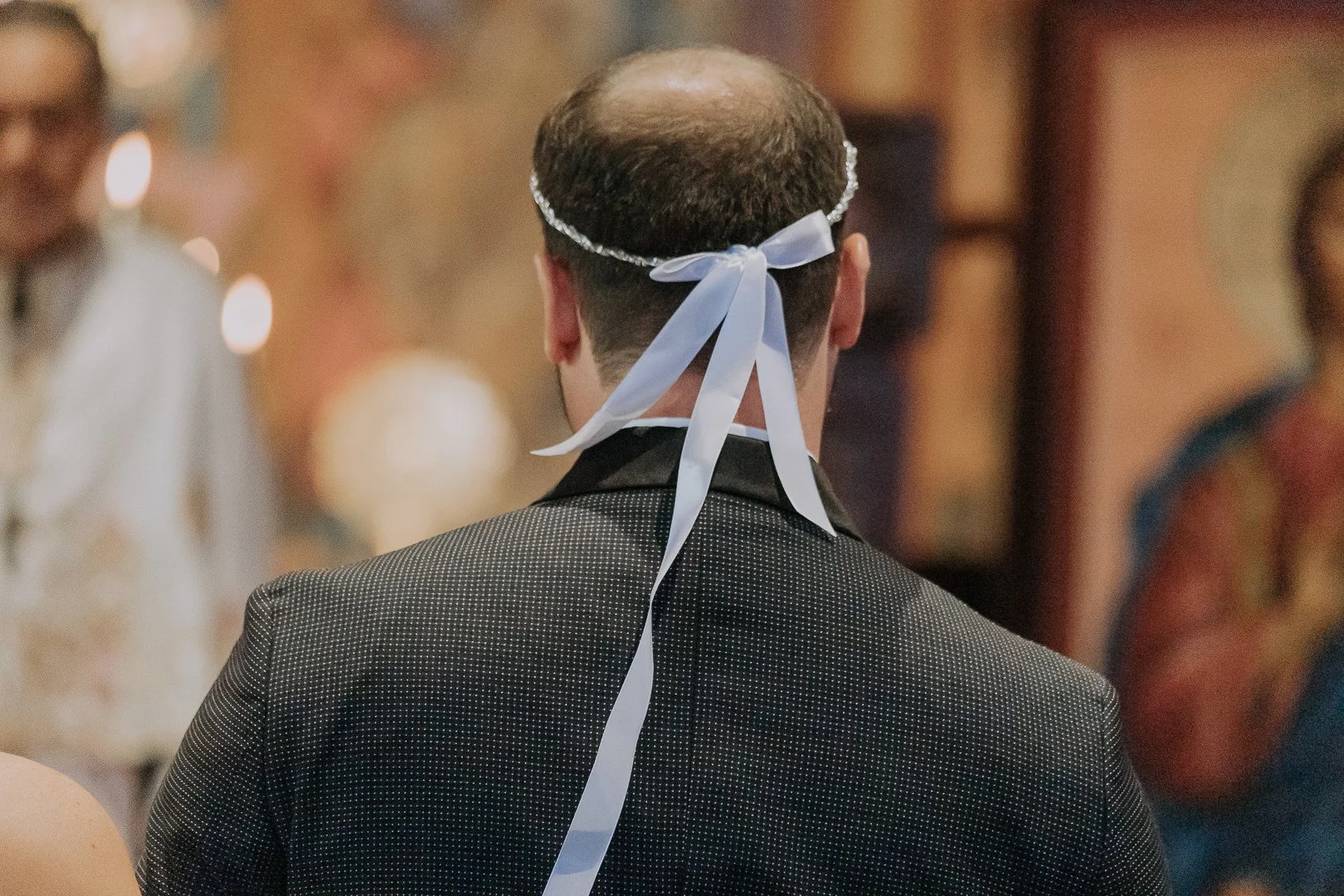 A person seen from behind wearing a dark dotted suit, with a white ribbon tied around the head in a bow, at an indoor event with blurred guests in the background.