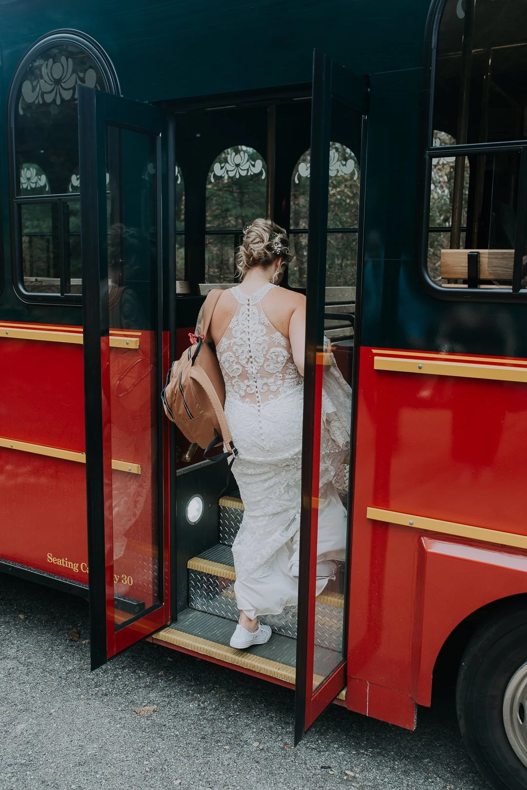 A woman in a white lace wedding dress with an intricate back design and a backpack, boarding a red and black trolley or sightseeing bus.