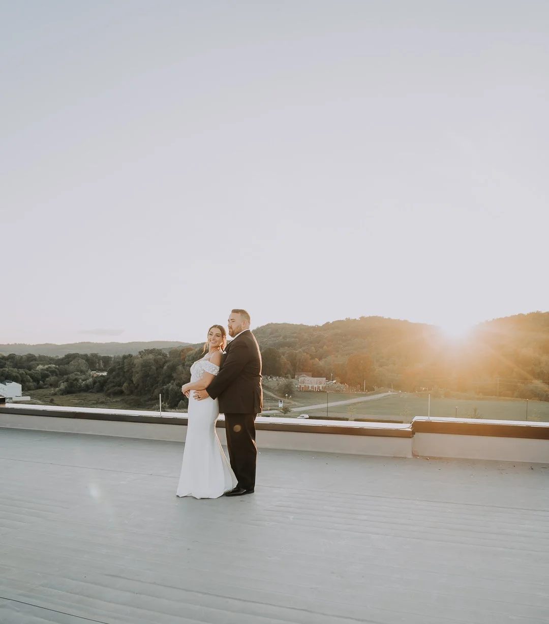 A bride and groom stand together on a rooftop during sunset, with the bride in a white wedding dress and the groom in a black suit, holding each other and smiling.