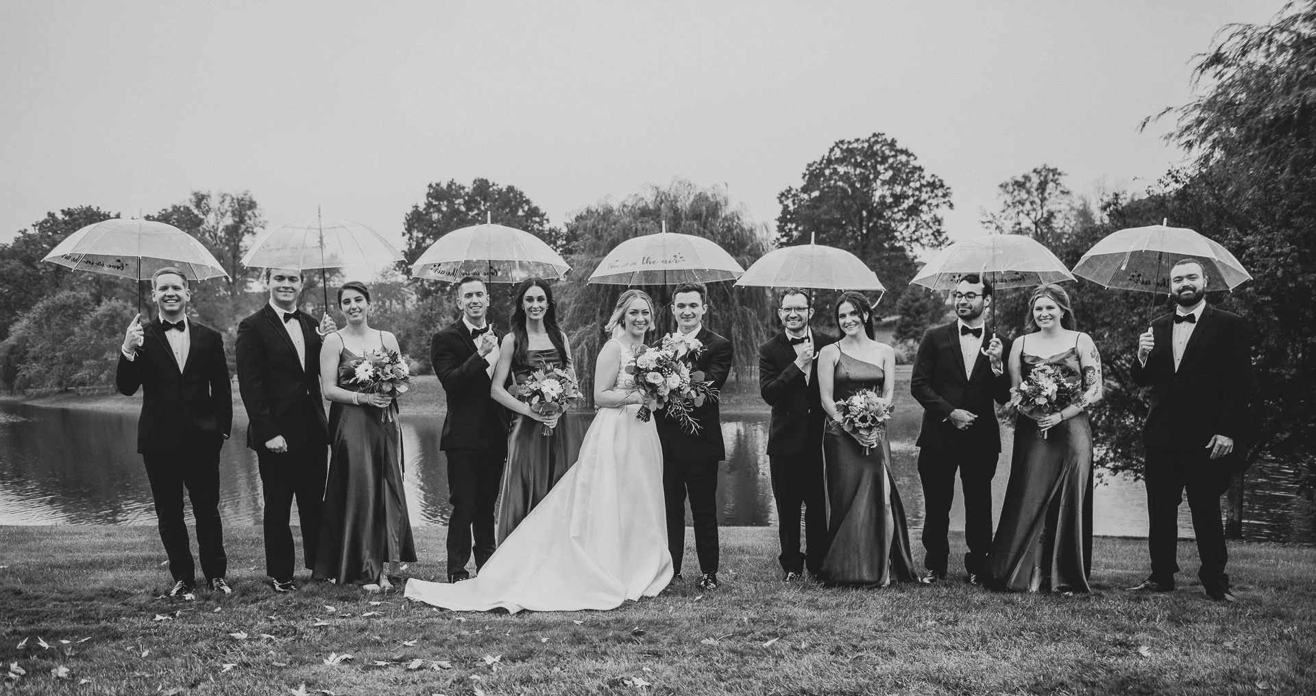 Black and white photo of a wedding party outdoors by a lake, with the bride and groom in the center, surrounded by bridesmaids and groomsmen holding umbrellas and bouquets.