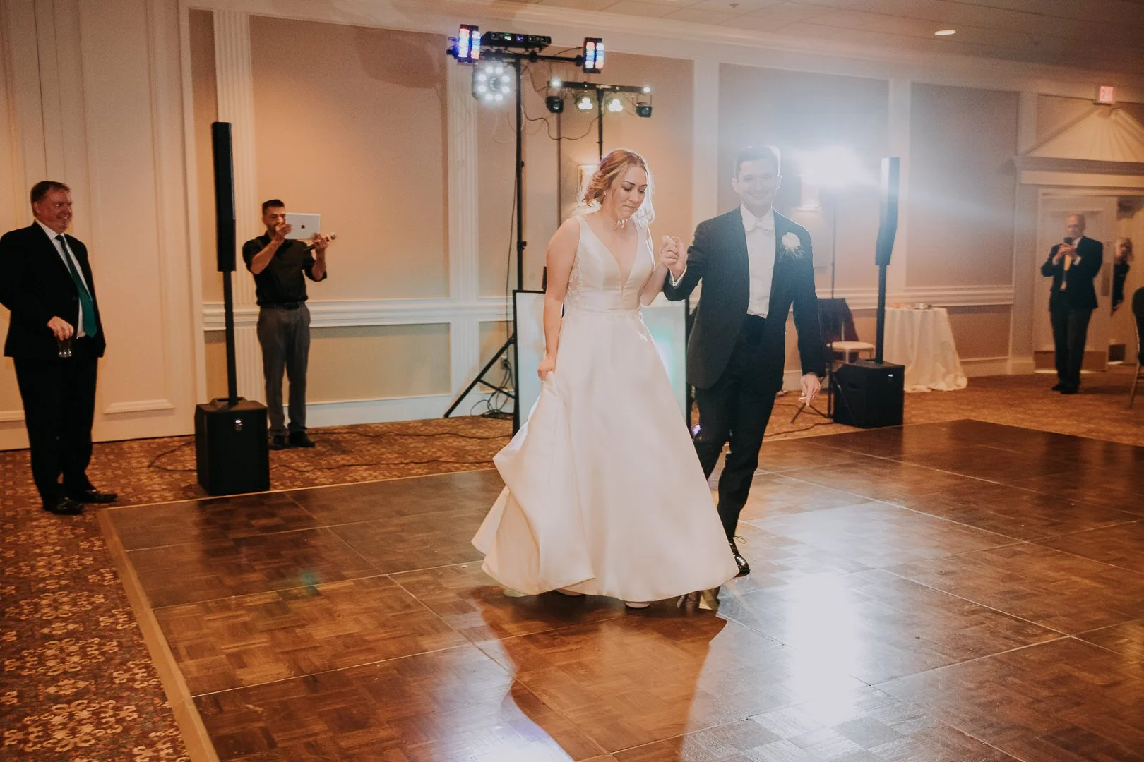 A bride and groom dancing at their wedding reception on a wooden dance floor, with guests and event staff watching and taking photos in the background, all indoors.
