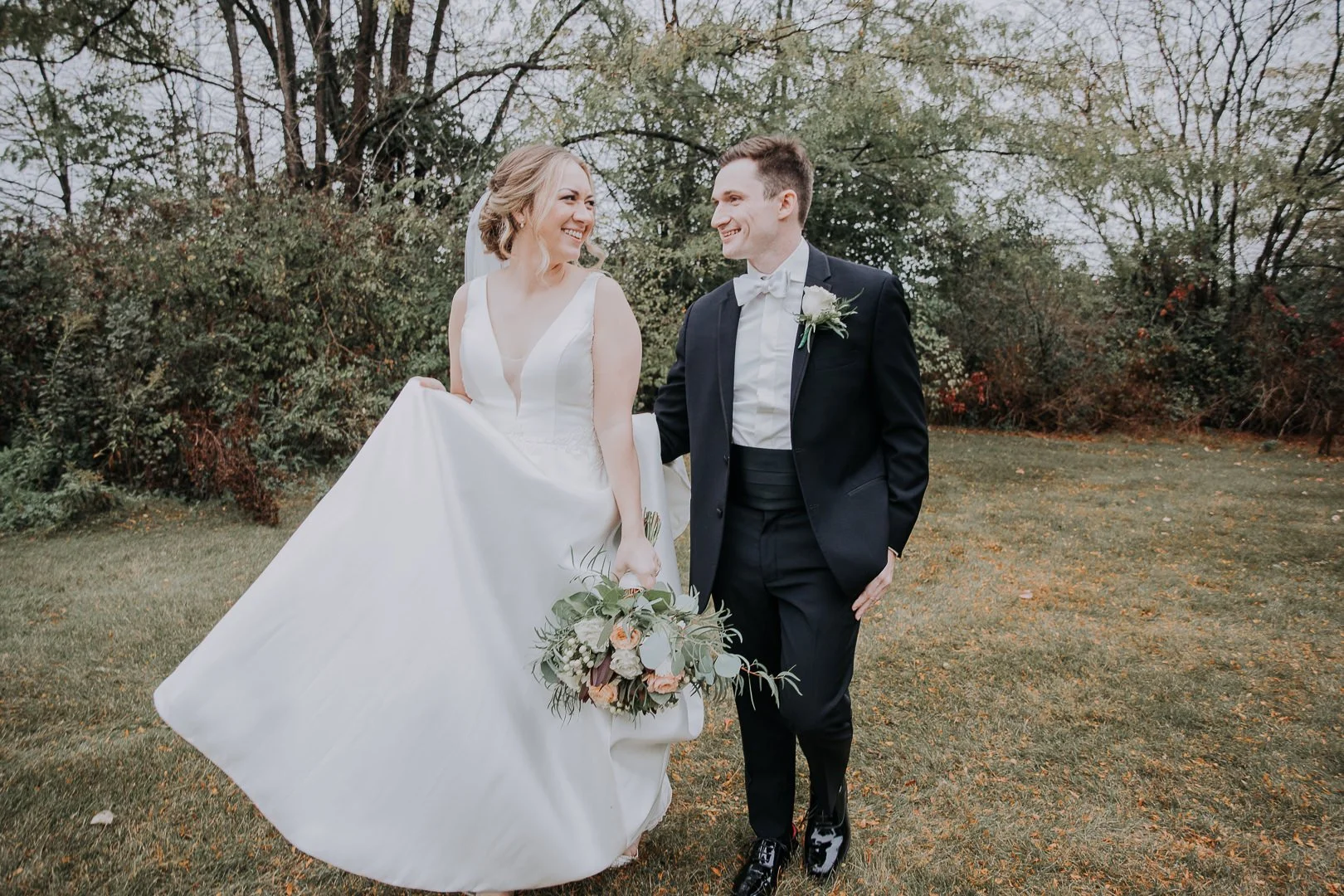 Bride and groom walking outside on their wedding day, smiling at each other, the bride holding her wedding dress and bouquet, the groom in a tuxedo with a white boutonniere.