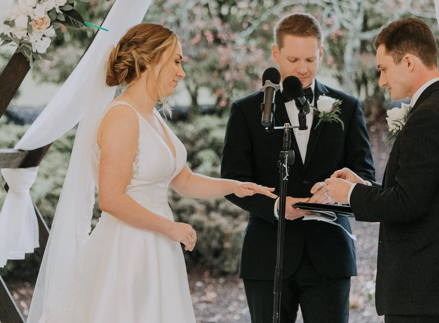 A bride and groom exchanging wedding vows under a wooden arch decorated with white fabric and flowers, with an officiant standing between them during an outdoor wedding ceremony.