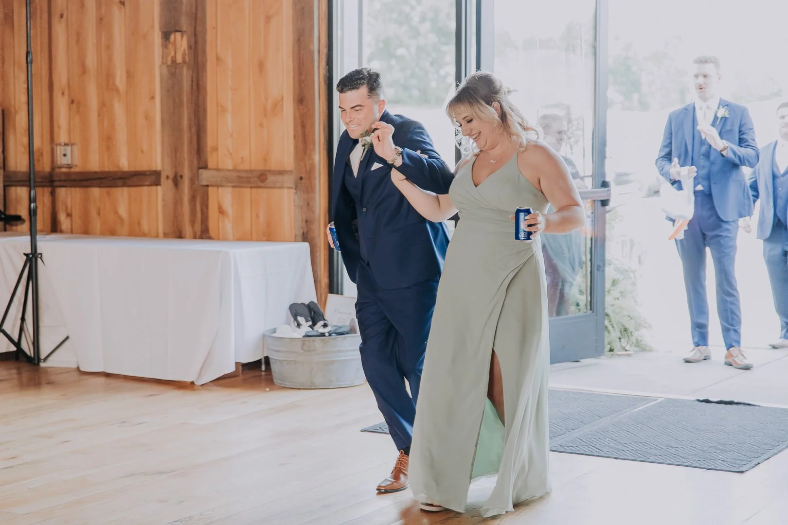 Groom and bride dancing at wedding reception, holding hands and smiling, with guests in the background near glass door