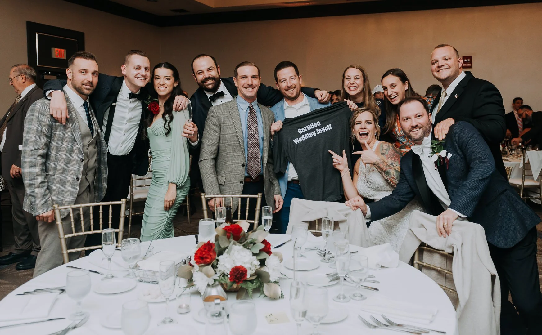 Group of people celebrating at a wedding reception, with some holding a T-shirt that says 'Certified Wedding Jagoff.'