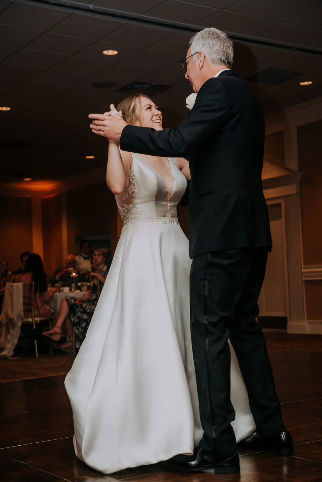 A bride and an older man dancing at a wedding reception, with guests seated in the background.