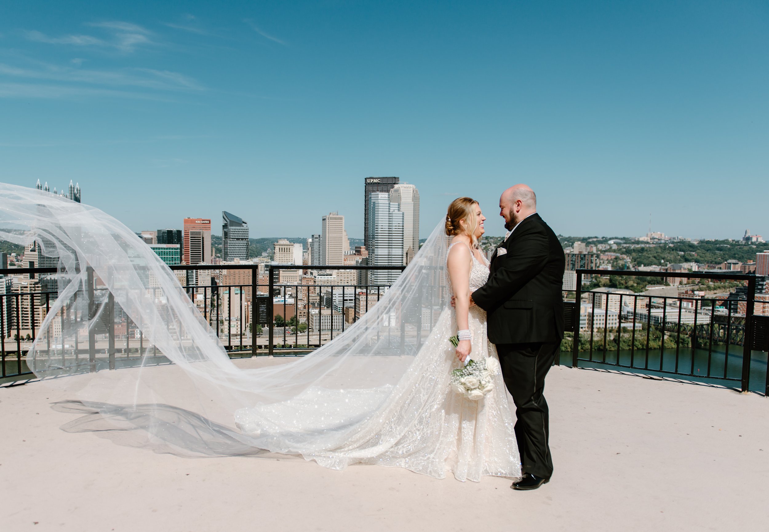 A bride and groom standing close to each other on a rooftop with a city skyline in the background. The bride is wearing a white wedding gown with a long veil, holding a bouquet of white flowers. The groom is dressed in a black suit, holding the bride