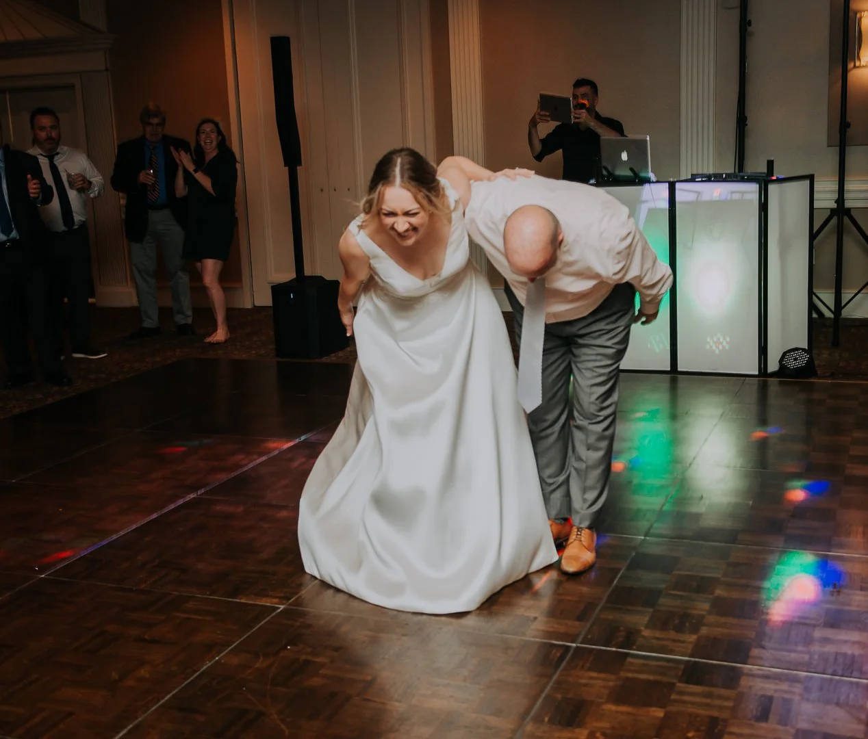 A woman in a white wedding dress laughs while a man in a light-colored shirt and gray pants, with a tie, leans over her, both bent at the waist, in a lively dance at a wedding reception. In the background, guests watch and take photos, and a DJ stand