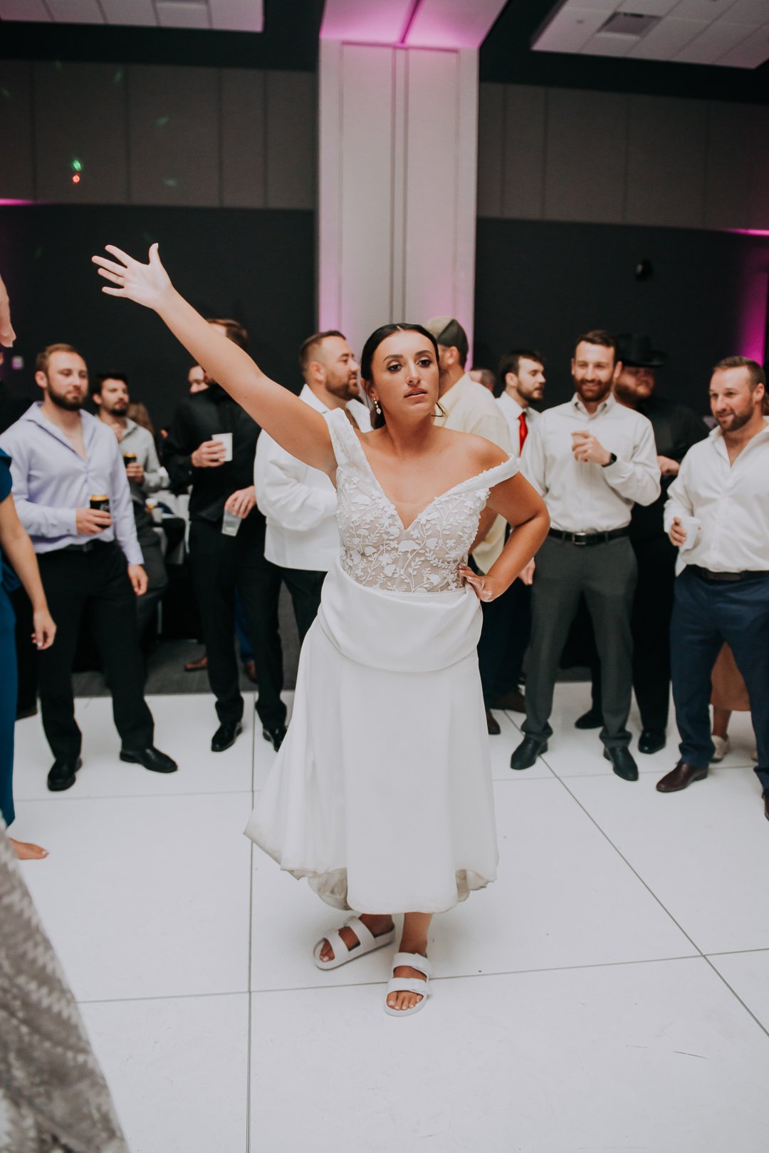 A woman in a wedding dress with one arm raised, standing on a dance floor at a wedding reception, surrounded by guests holding drinks.