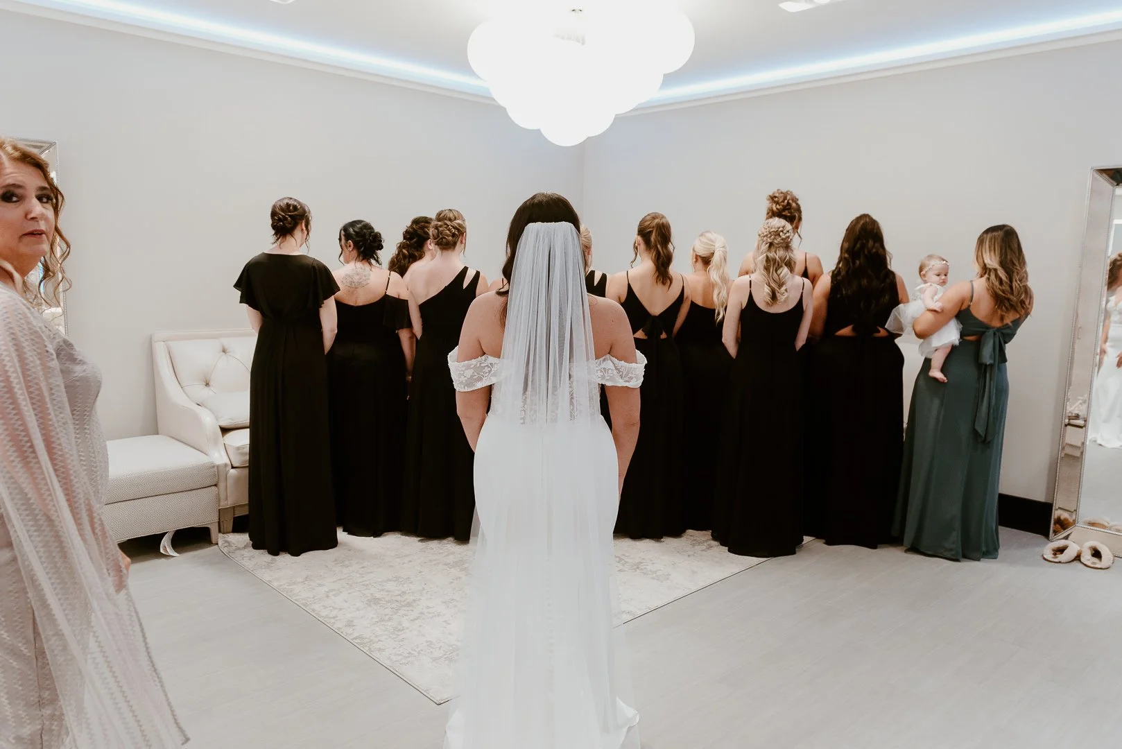Bride in white wedding dress with veil standing with back to camera, facing a group of women dressed in black, inside a modern, well-lit room.