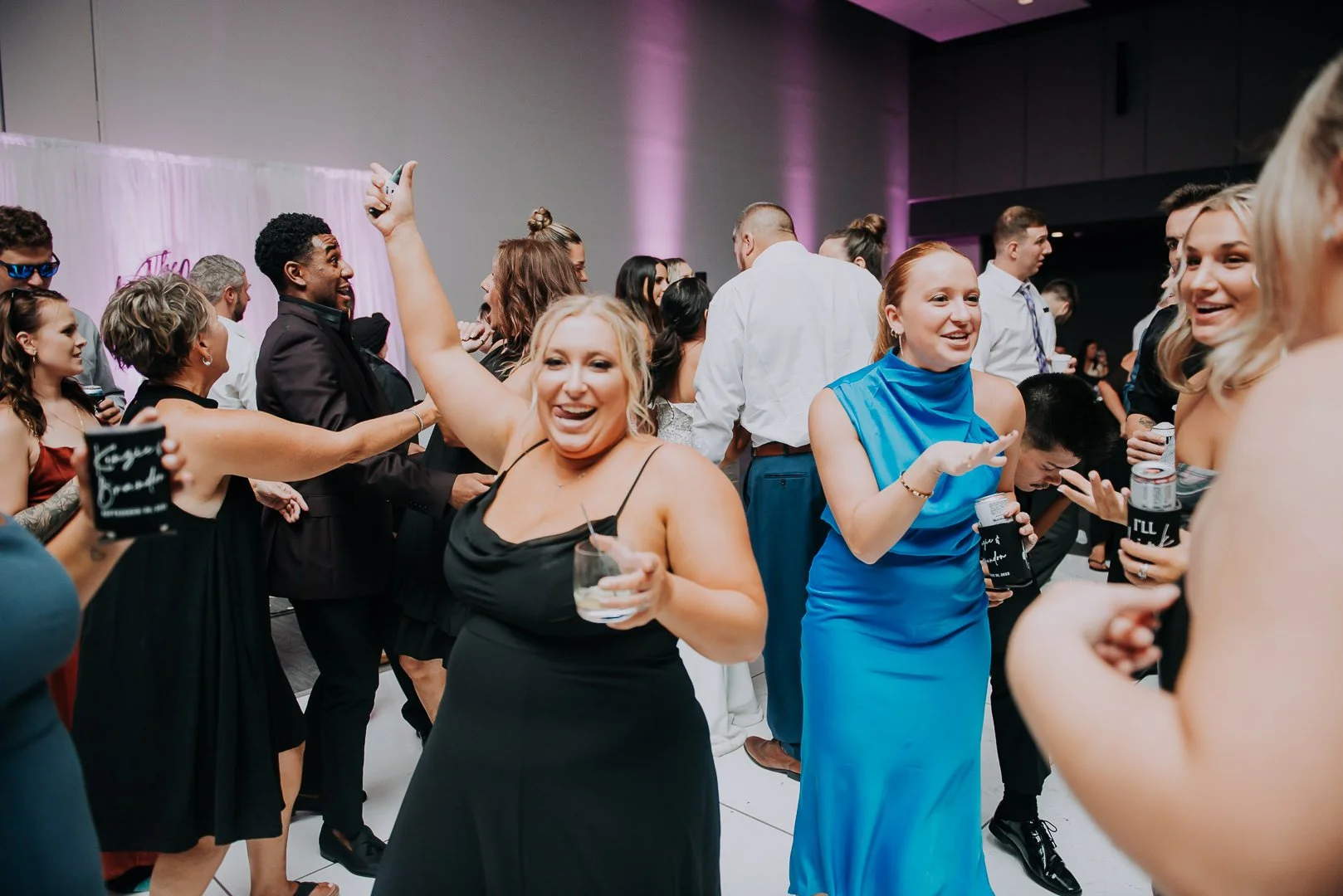 People dancing and celebrating at a party, with women holding drinks and smiling, wearing formal attire.