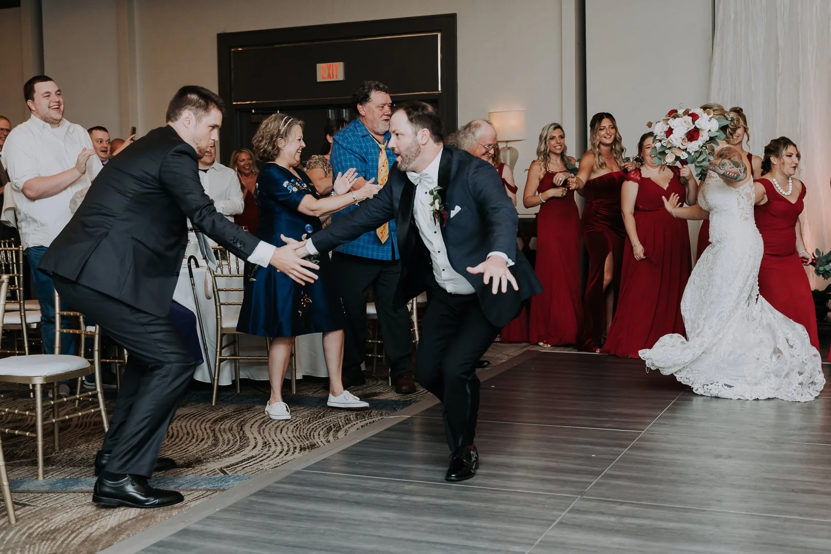 Two men in tuxedos dancing at a wedding reception, with a bride holding a large bouquet and women in red dresses in the background, some clapping and smiling.