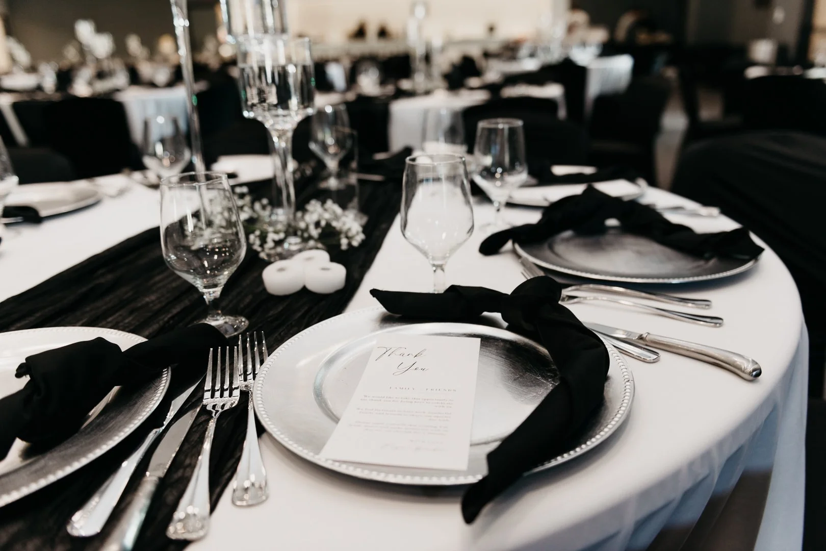 Elegant banquet table set with silverware, black napkins tied with a knot, clear glassware, silver chargers, white plates, and a black table runner with white candles and flowers, ready for a formal event or wedding reception.