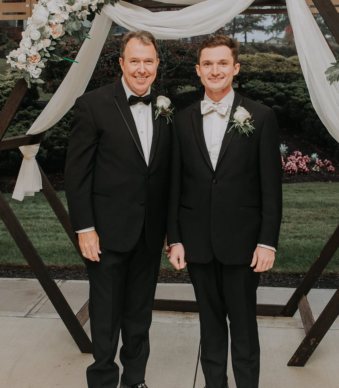 Two men in black tuxedos with white shirts and black bow ties, standing side by side outdoors in front of a decorated wooden archway, smiling at the camera. One man has a white boutonniere on his lapel, and the other has a similar boutonniere with a 