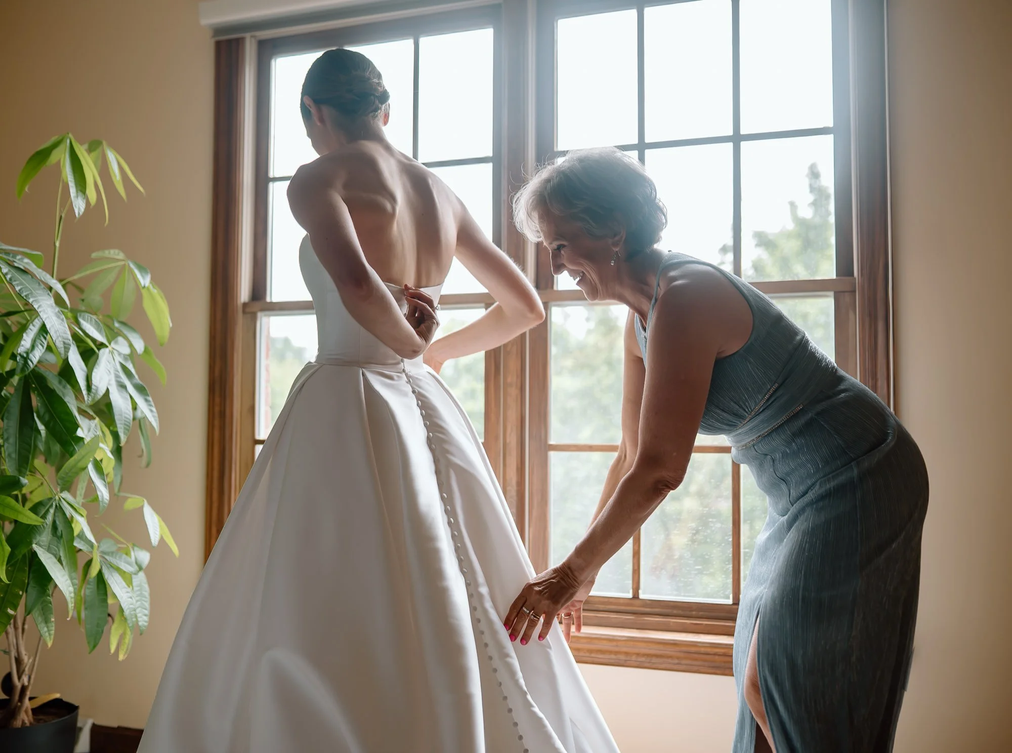 A senior woman in a gray dress helping a young woman in a wedding dress adjust the back of her gown by a window with sunlight streaming inside.