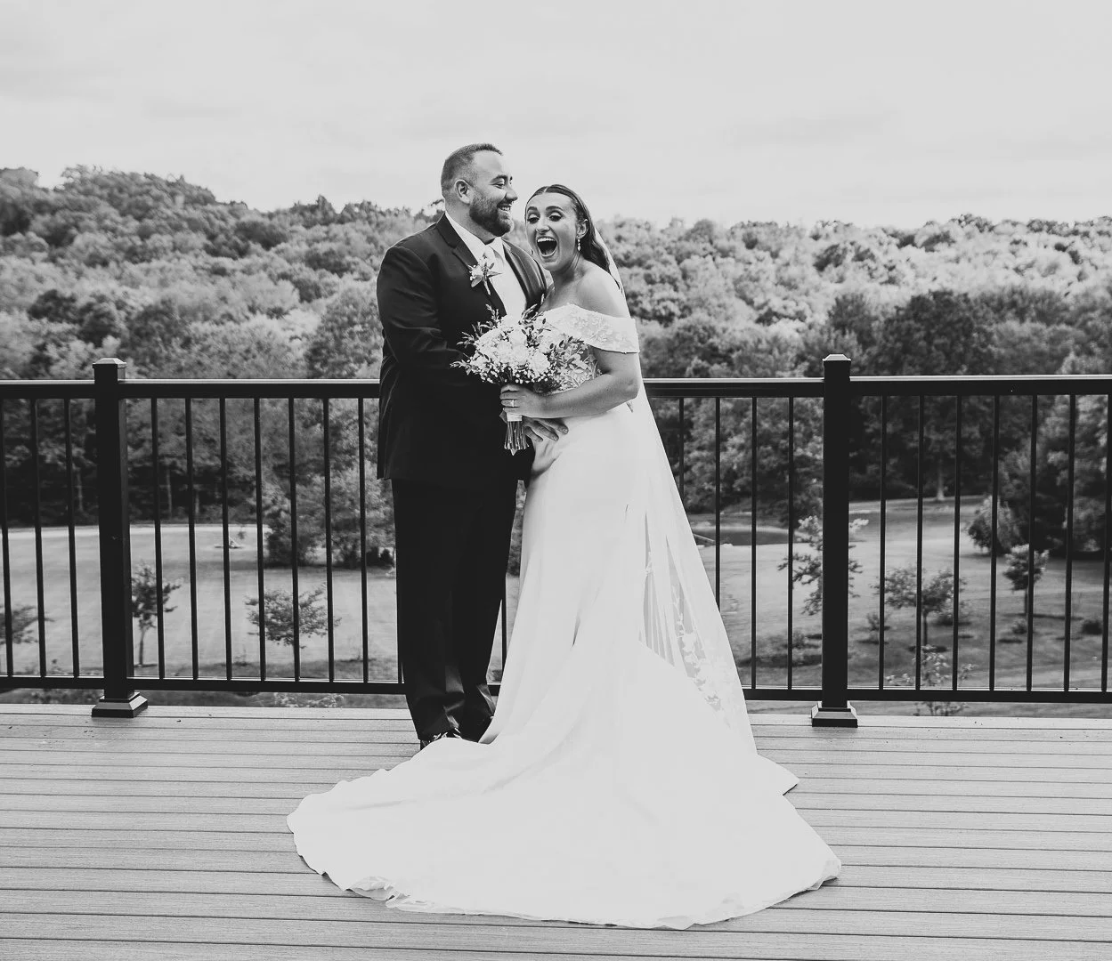 Black and white photo of a newlywed couple on a balcony, smiling and hugging, with a scenic forest in the background. The bride is holding a bouquet of flowers, wearing an off-shoulder wedding gown, and the groom is in a suit.