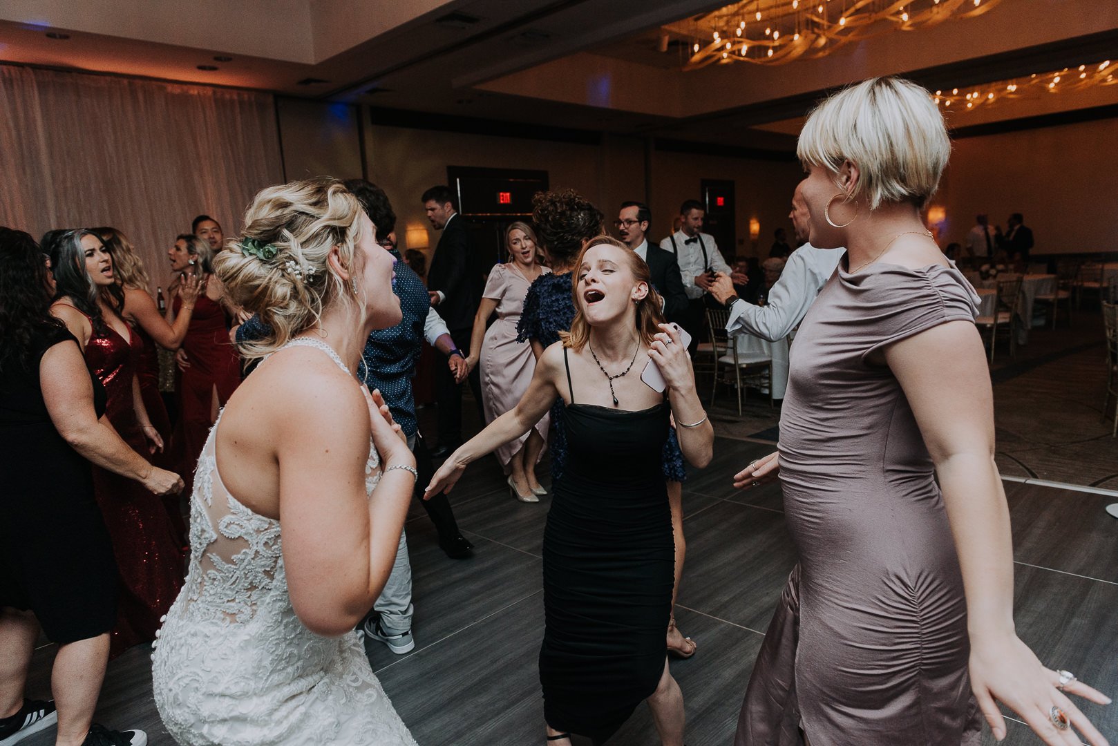 People dancing and celebrating at a wedding reception, with women in elegant dresses and guests enjoying the music in a decorated event hall.