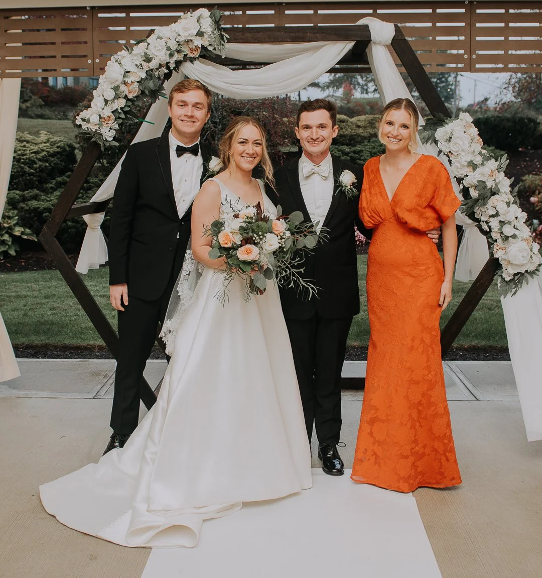 Group of five people at a wedding, standing under a wooden arch decorated with white flowers and draped fabric, outdoors on a garden lawn.