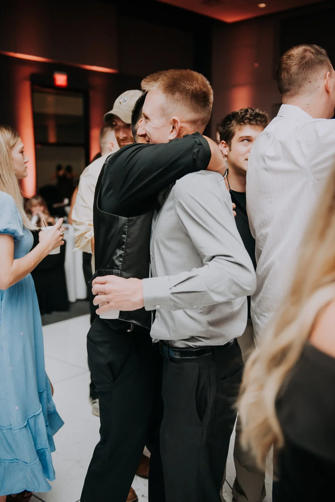 Two young men hugging at a social event, surrounded by other people.