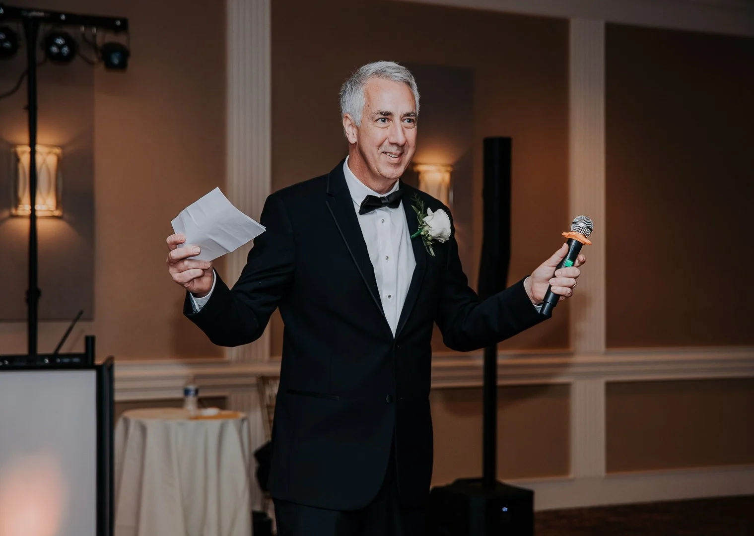 A man in a tuxedo with a white boutonniere giving a speech at an indoor event, holding a piece of paper in one hand and a microphone in the other.