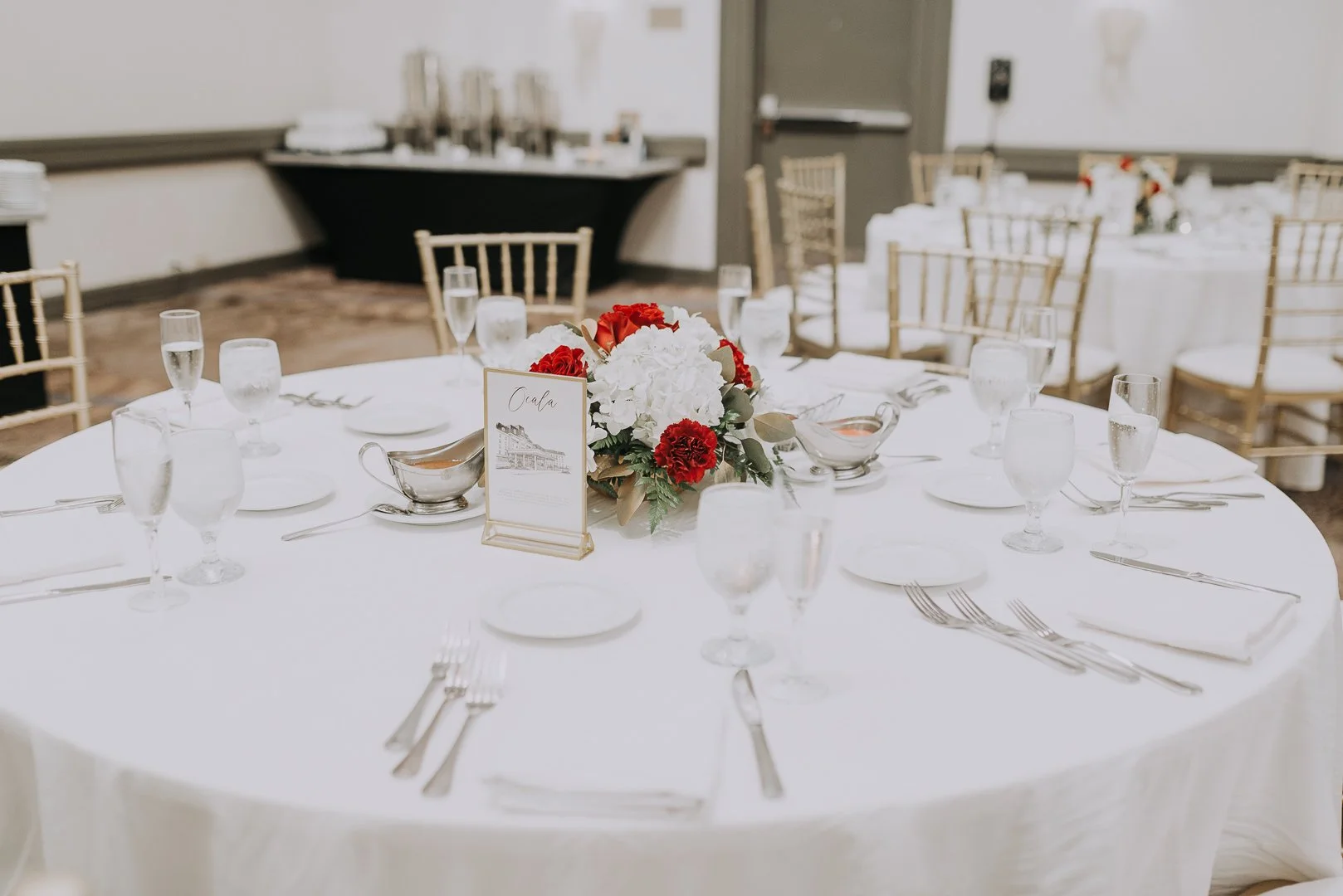 Round banquet table set for a formal event with a floral centerpiece of white and red flowers, water glasses, white plates, and silverware, with chairs arranged around it in an elegant venue.