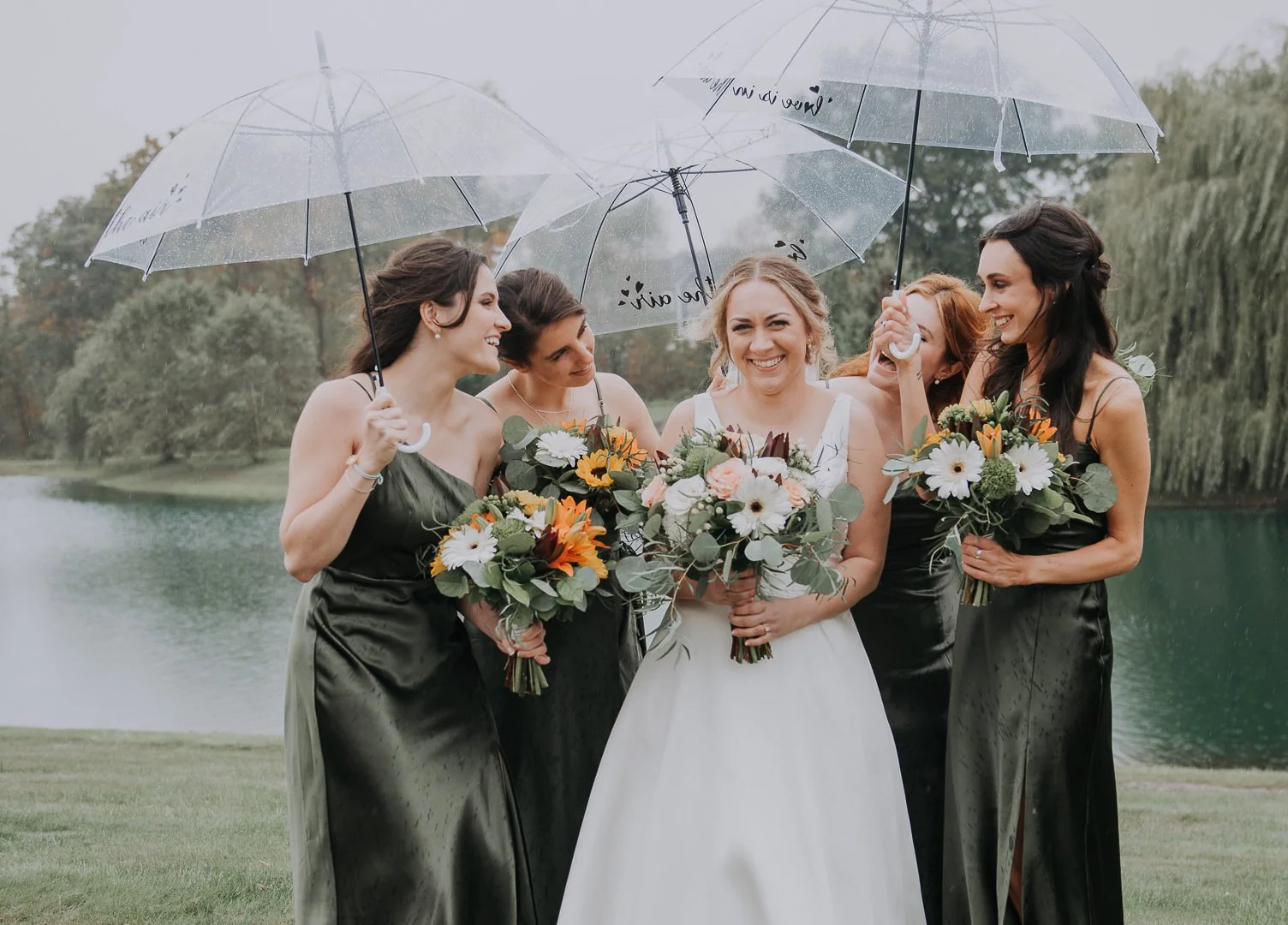 A bride in a white wedding dress holding a bouquet of flowers surrounded by four women in black dresses holding umbrellas and bouquets, standing by a lake with trees in the background on a rainy day.