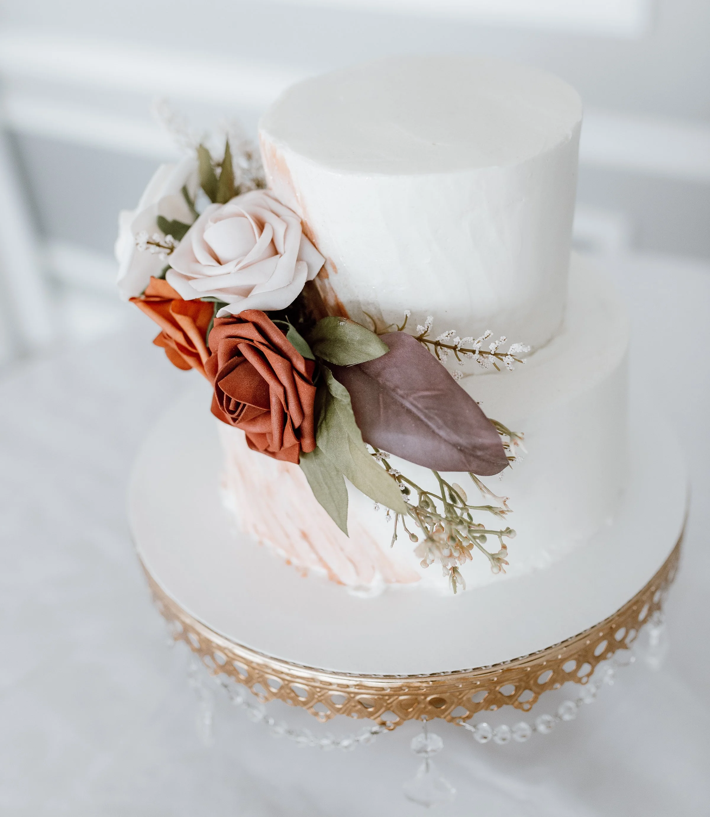 A two-tiered white wedding cake decorated with a bouquet of faux roses and greenery on the side, placed on a decorative cake stand.