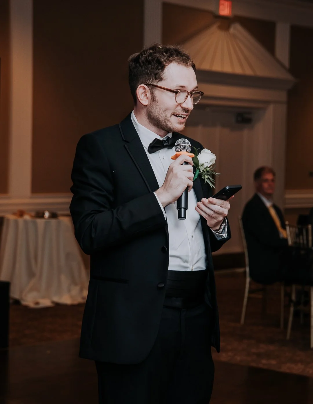 A man in a tuxedo with a white boutonniere speaking into a microphone while looking at his phone at an event.