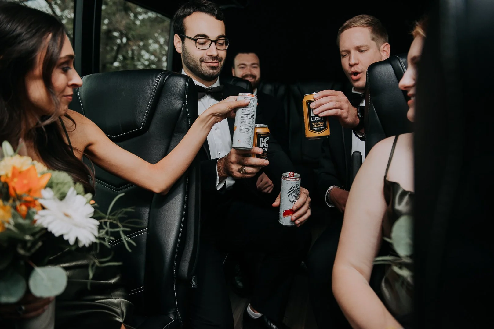 Group of young adults wearing formal attire, some holding canned beverages, sitting in the back of a vehicle, possibly celebrating or socializing.