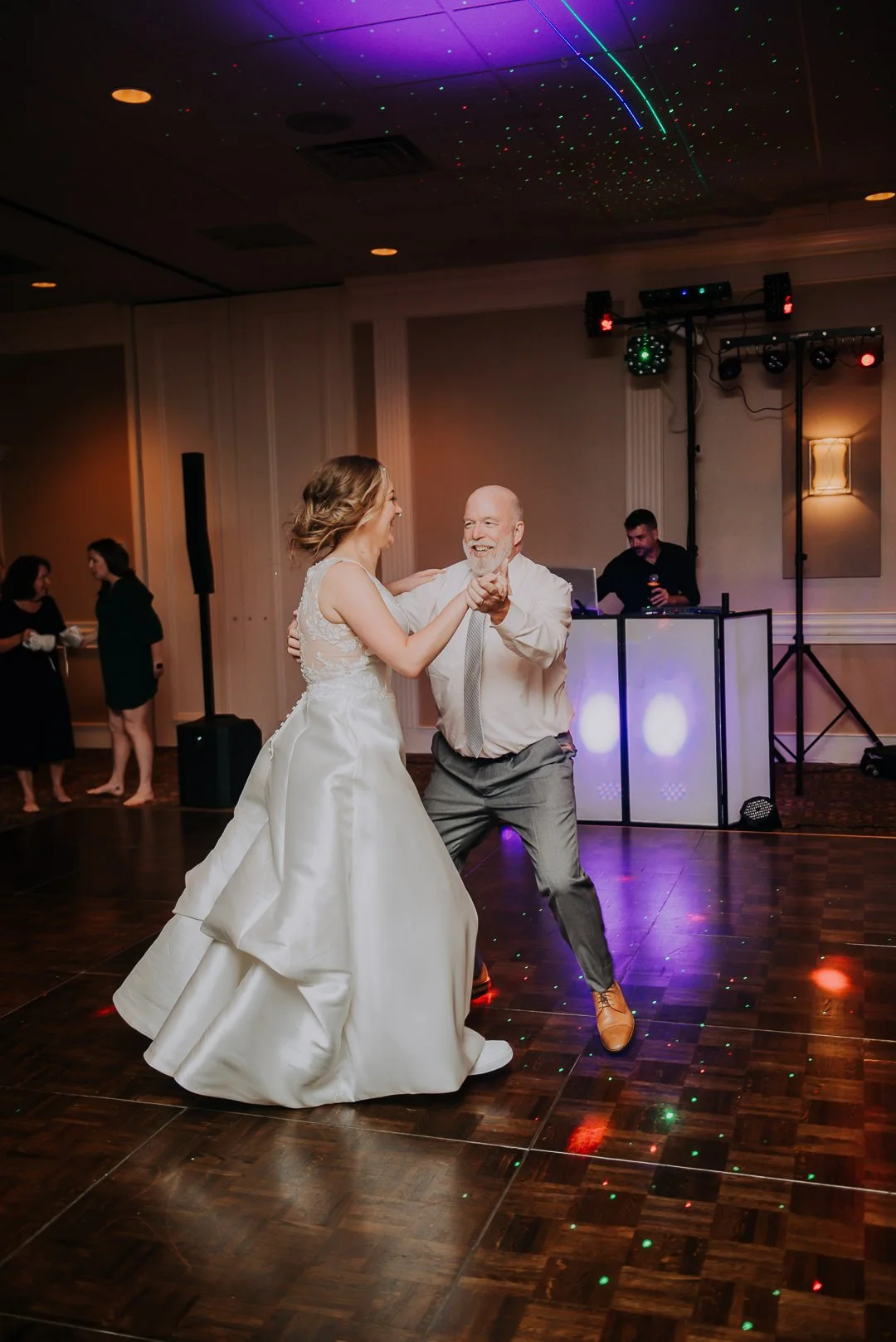 A bride and an older man dancing at a wedding reception, with a DJ in the background and colorful lights on the ceiling and dance floor.