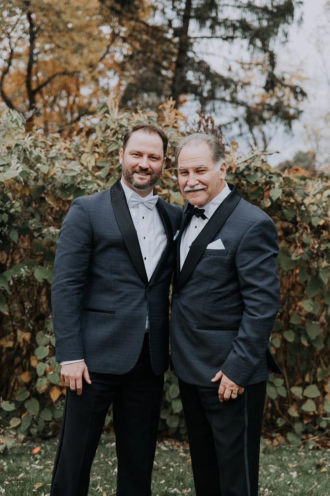 Two men in tuxedos standing together outdoors among autumn foliage, smiling at the camera.