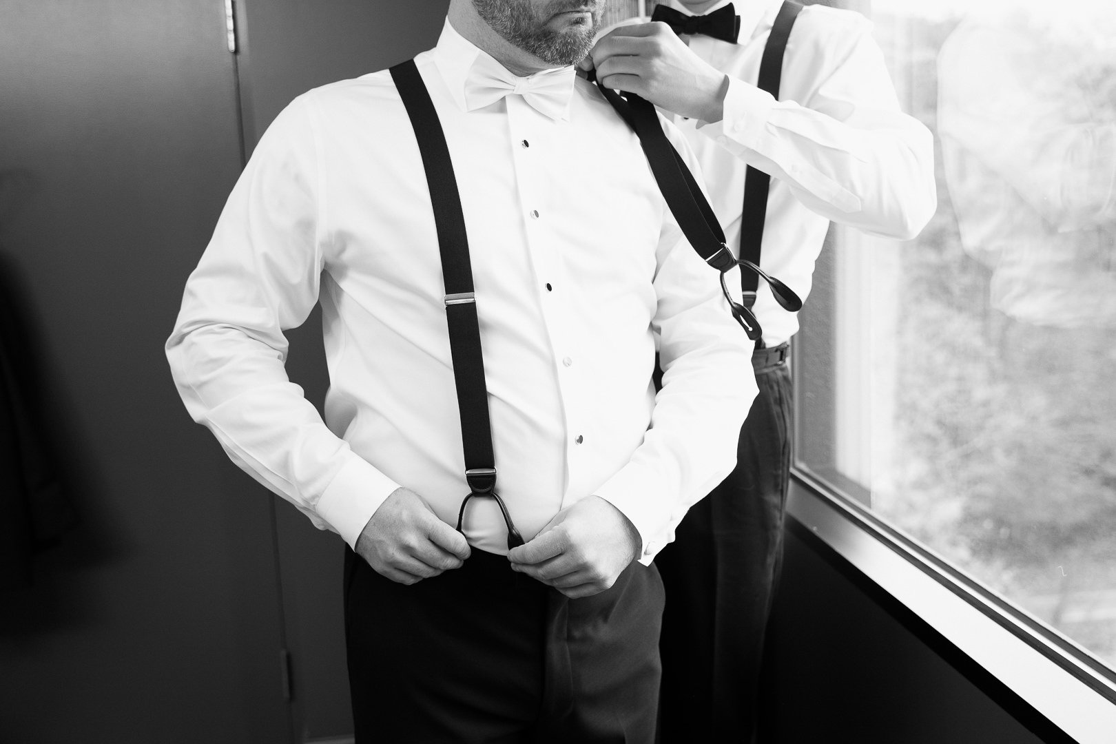 Man adjusting suspenders in a formal outfit, with another man helping with a bow tie, near a window in black and white.