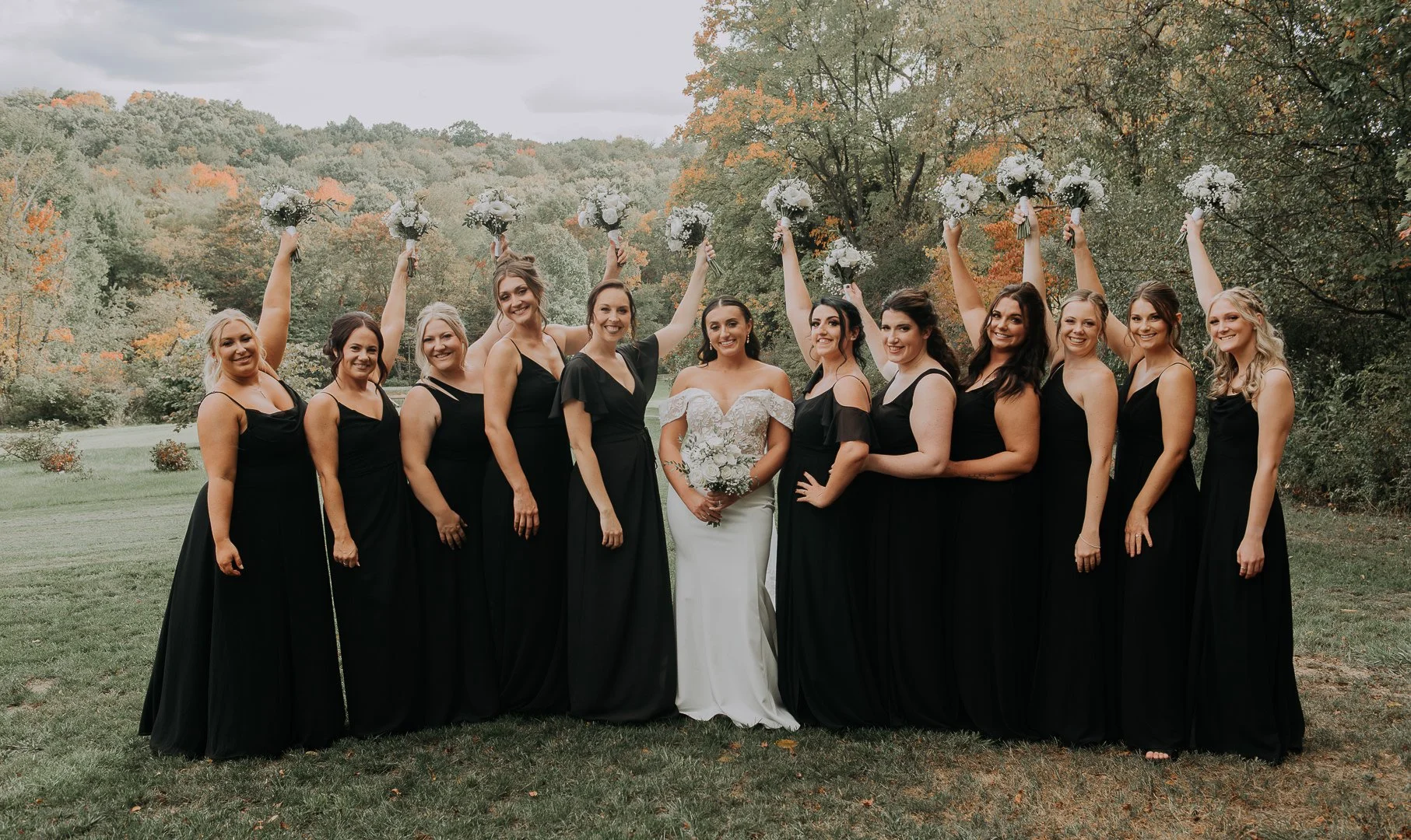 Bride and bridesmaids standing outdoors in a grassy field with autumn trees in the background. The bride is in a white dress holding a bouquet, surrounded by women in black dresses, some holding bouquets raised in the air.