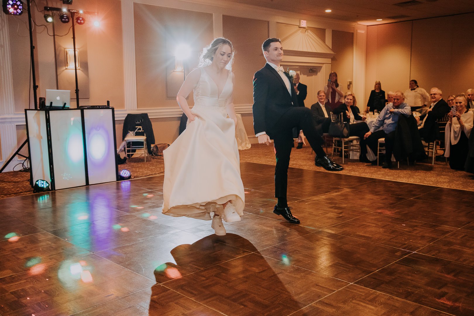 Bride and groom dancing on a wooden floor in a wedding reception hall, with guests seated at tables watching. The bride wears a white wedding gown, and the groom wears a black tuxedo. There are colorful lights and a DJ setup nearby.