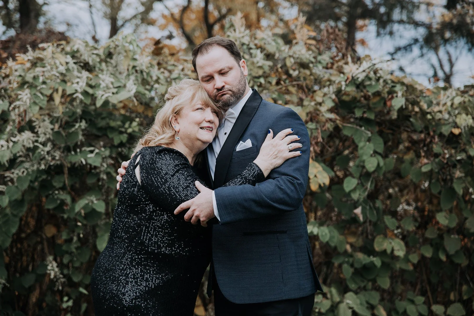 A man and an older woman hugging each other outdoors in front of green foliage. The woman is smiling, and the man has a serious expression. They are dressed in formal attire.